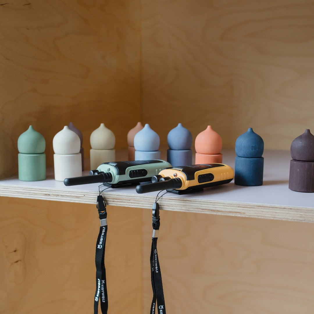 Green and orange KIDYTALK walkie-talkies on a wooden shelf with their black Kidywolf lanyards hanging down