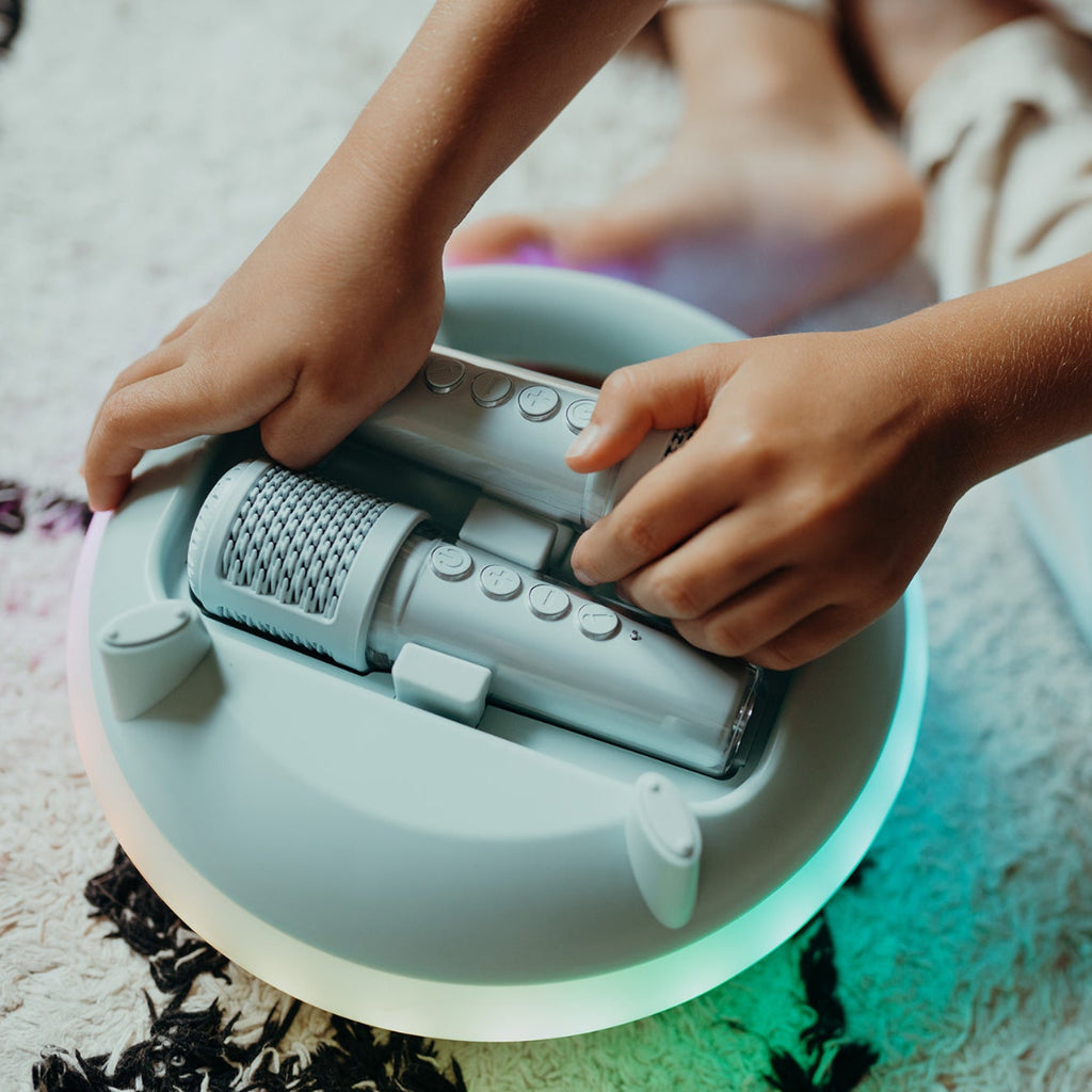 A close-up of a child's hands placing a light blue microphone into a karaoke machine white textured rug. The toy glows with a soft green light from underneath.