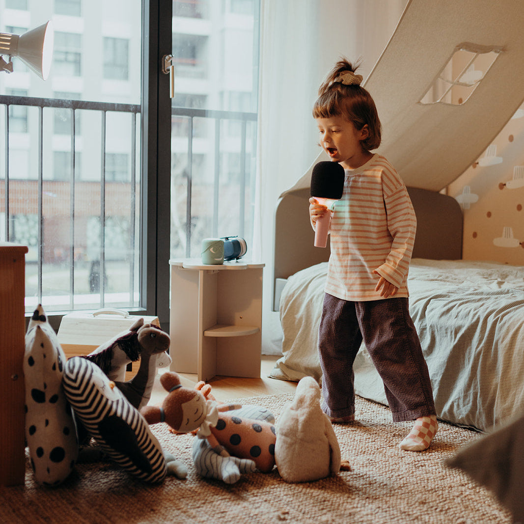 Child holding a microphone in a room with toys and large windows.