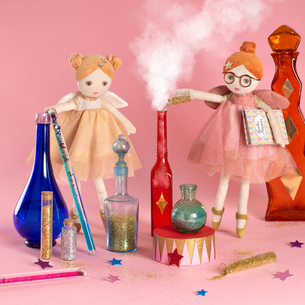 Two dolls with a pink background, surrounded by decorative bottles and items.
