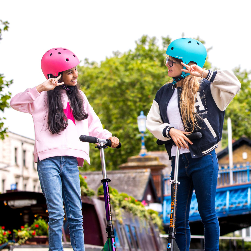 Two girls on a city bridge with scooters, one wearing a pink Micro helmet and one fastening an aqua Micro helmet