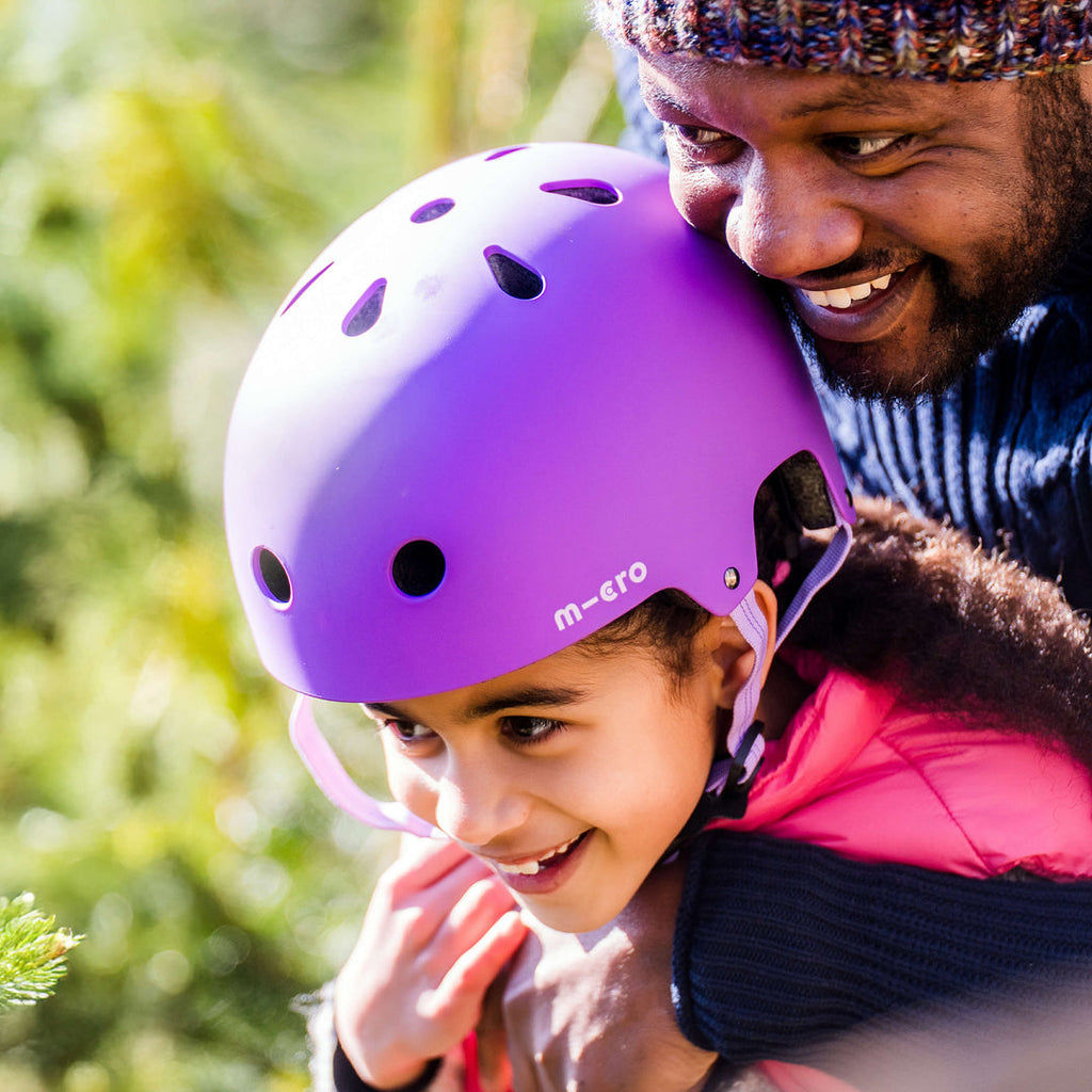 Smiling child wearing the purple Micro helmet being hugged by a laughing adult outdoors