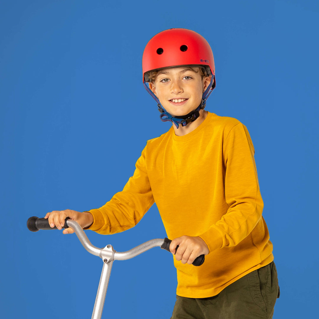 Smiling boy in a mustard jumper wearing the red Micro helmet and holding scooter handlebars against a blue background
