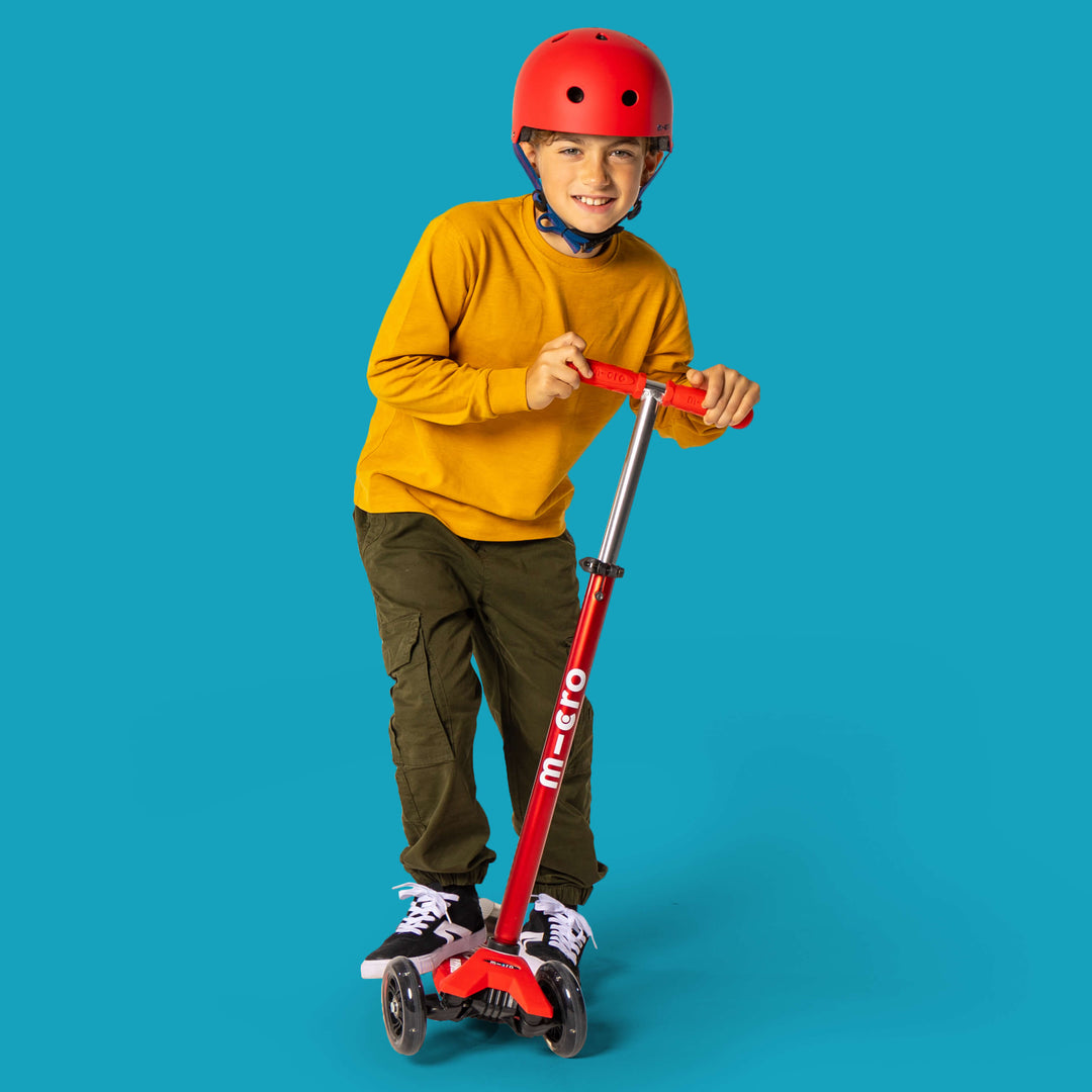 Boy wearing the red Micro helmet riding a red Micro scooter against a teal background