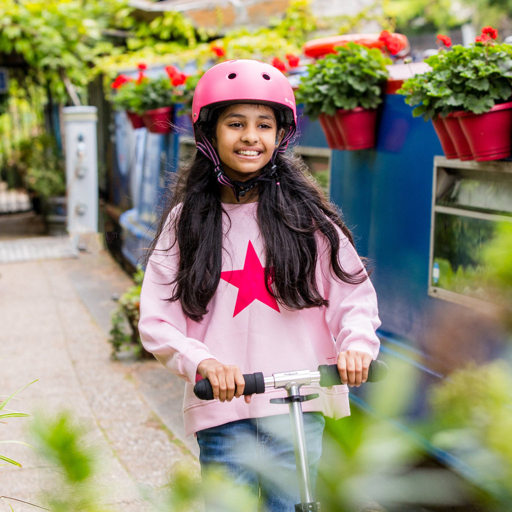 Smiling girl in a pink star jumper wearing the pink Micro Eco helmet while riding a scooter past flower boxes