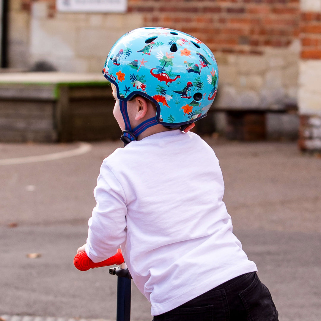 Child seen from behind wearing the Dino Micro helmet while riding a scooter in a playground
