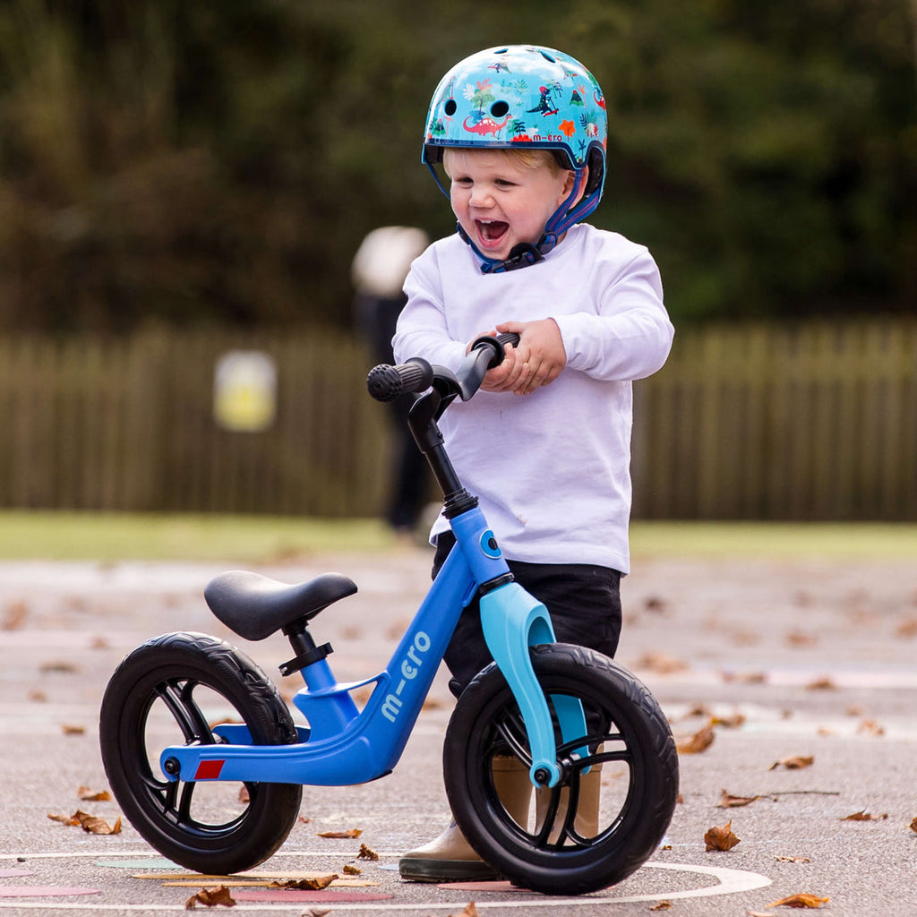 Laughing toddler wearing the Dino Micro helmet standing with a blue Micro balance bike