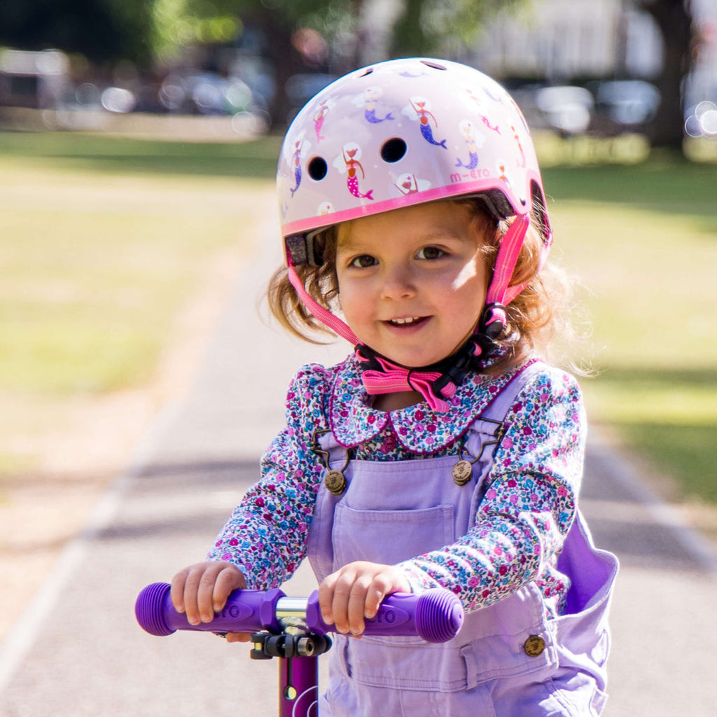Smiling toddler in purple dungarees wearing the Mermaid Micro helmet while riding a scooter in a park