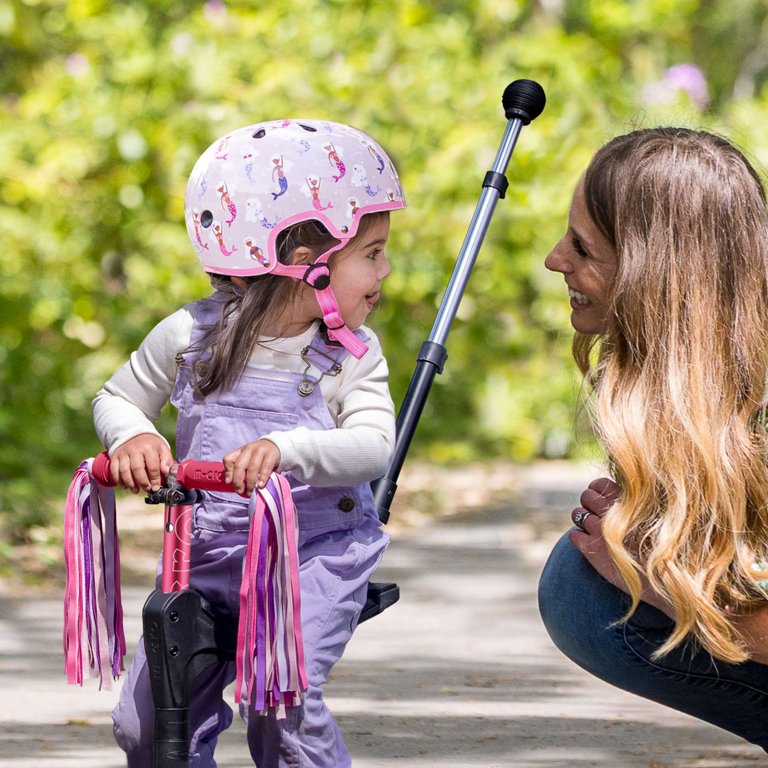 Toddler in the Mermaid Micro helmet on a ride-on scooter with pink ribbons, chatting to a parent