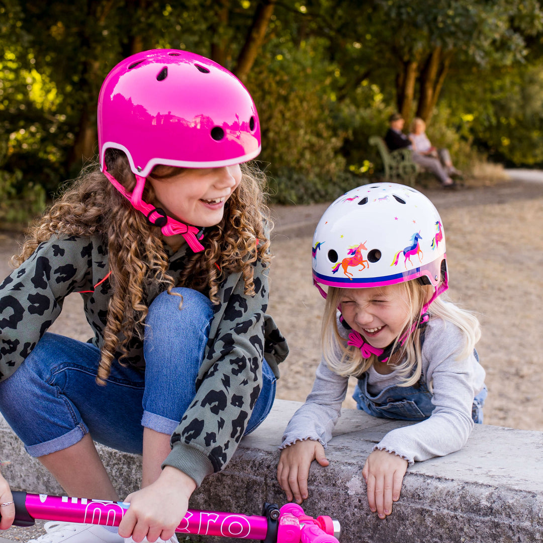 Two smiling girls in Micro helmets, one pink and one Unicorn, crouched beside a pink scooter in a park