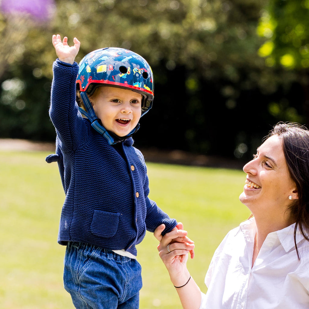 Smiling toddler in a navy cardigan wearing the Vehicle Micro helmet, holding hands with a laughing woman on a lawn