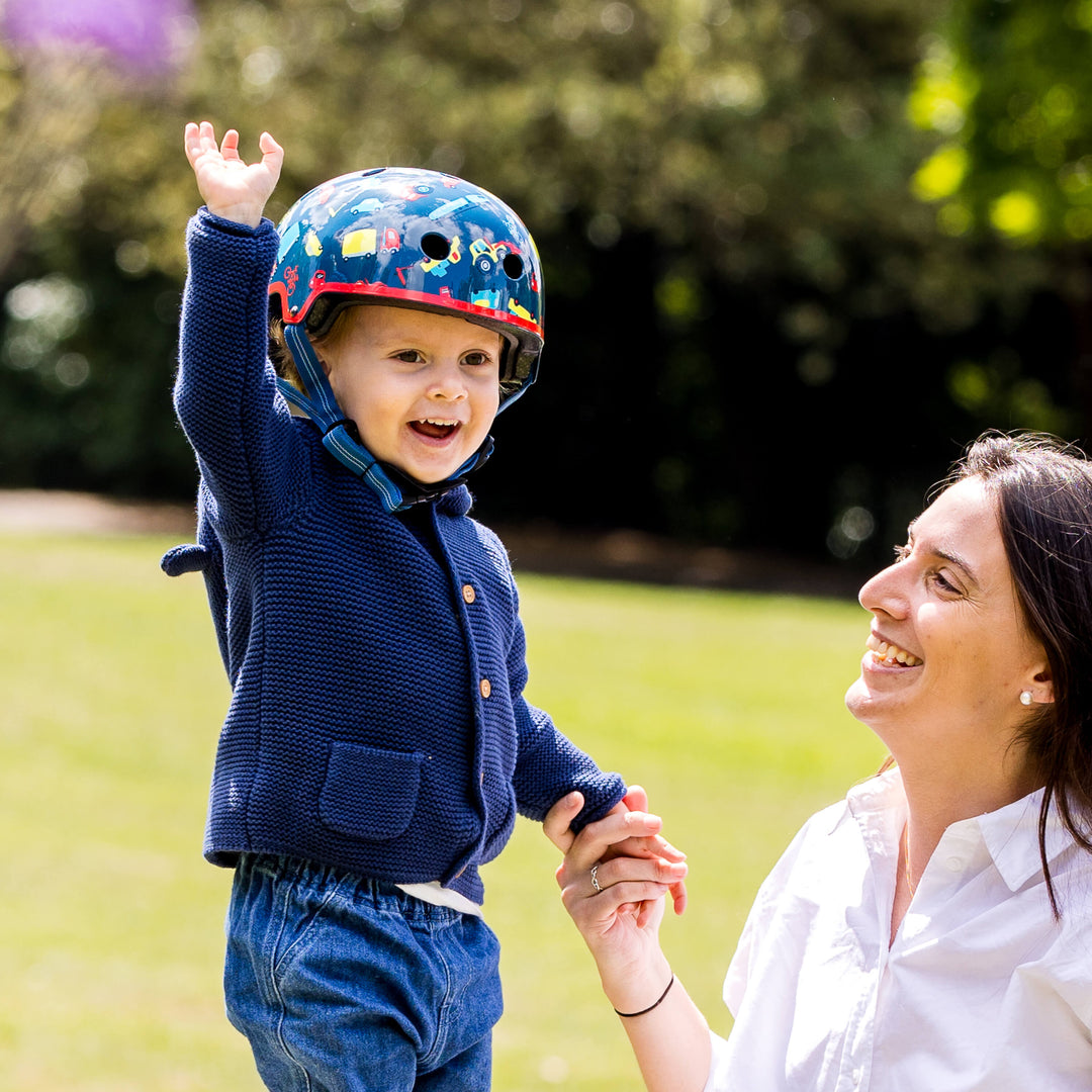 Smiling toddler in a navy cardigan wearing the Vehicle Micro helmet, holding hands with a laughing woman on a lawn