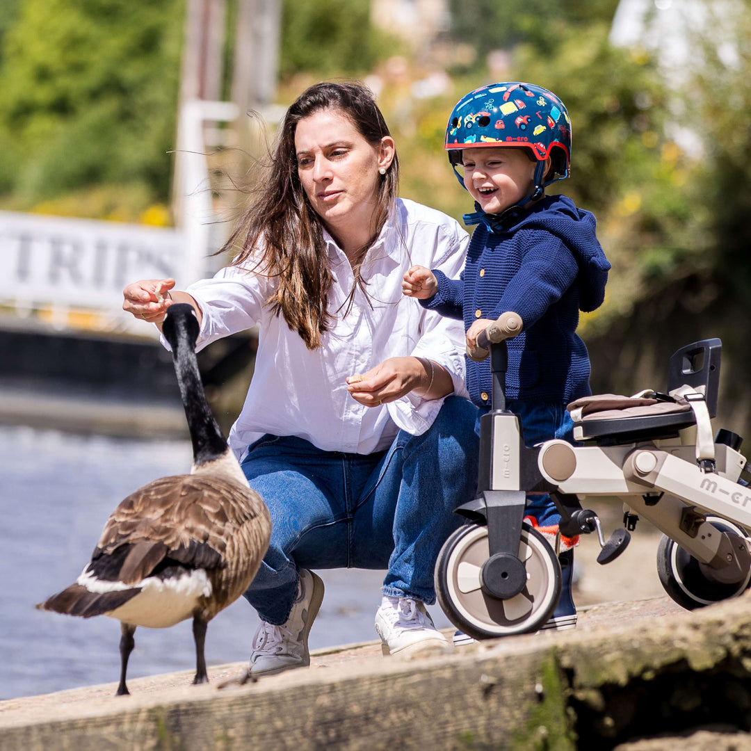 Woman and toddler in the Vehicle Micro helmet feeding a goose by a river, with a Micro ride-on beside them
