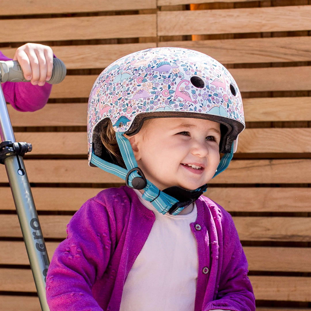 Smiling toddler in a purple cardigan wearing the Flora-Saur Micro helmet next to a scooter