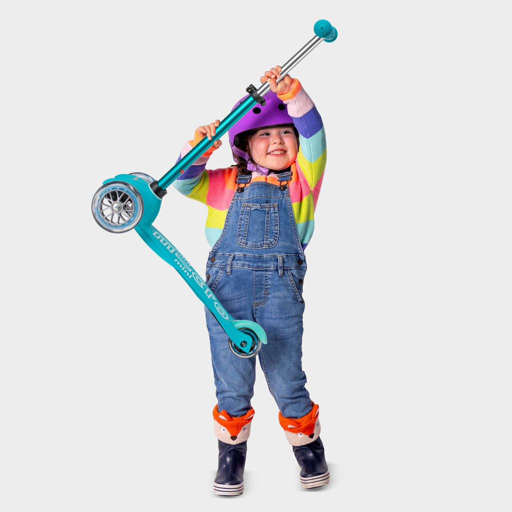 Toddler in a rainbow jumper and dungarees lifting the lightweight aqua Mini Micro Deluxe scooter overhead