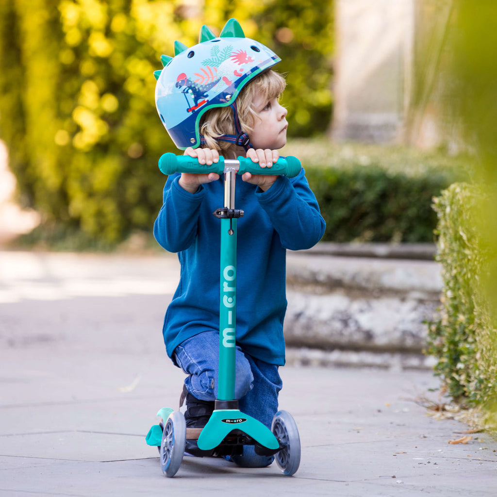 Toddler in a dinosaur helmet kneeling on the aqua Mini Micro Deluxe scooter on a garden path