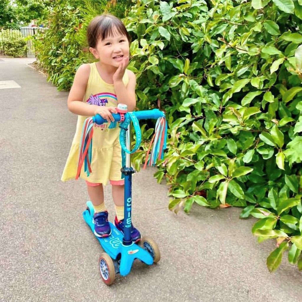 Smiling girl in a yellow dress standing on a blue Mini Micro scooter with ribbon streamers on a path