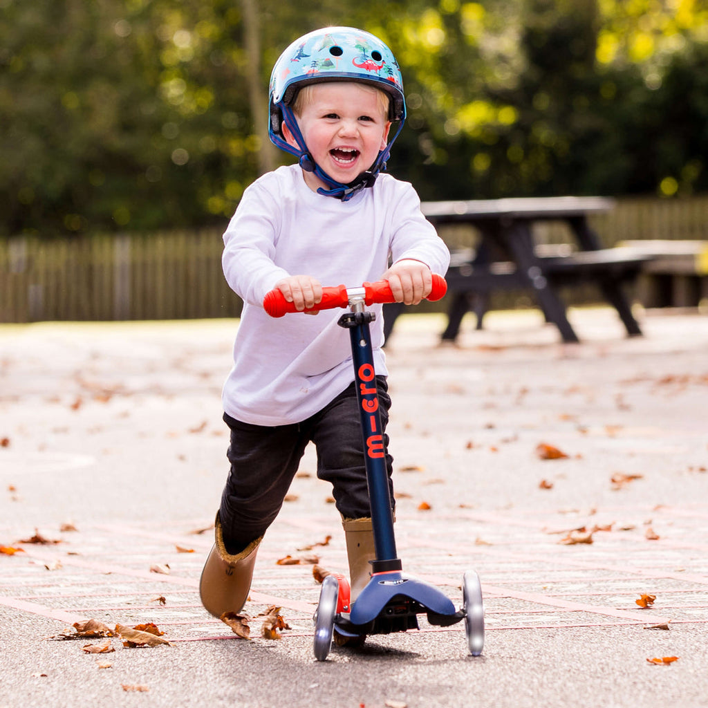 Laughing toddler in a helmet and white top riding the navy Mini Micro Deluxe scooter in a playground