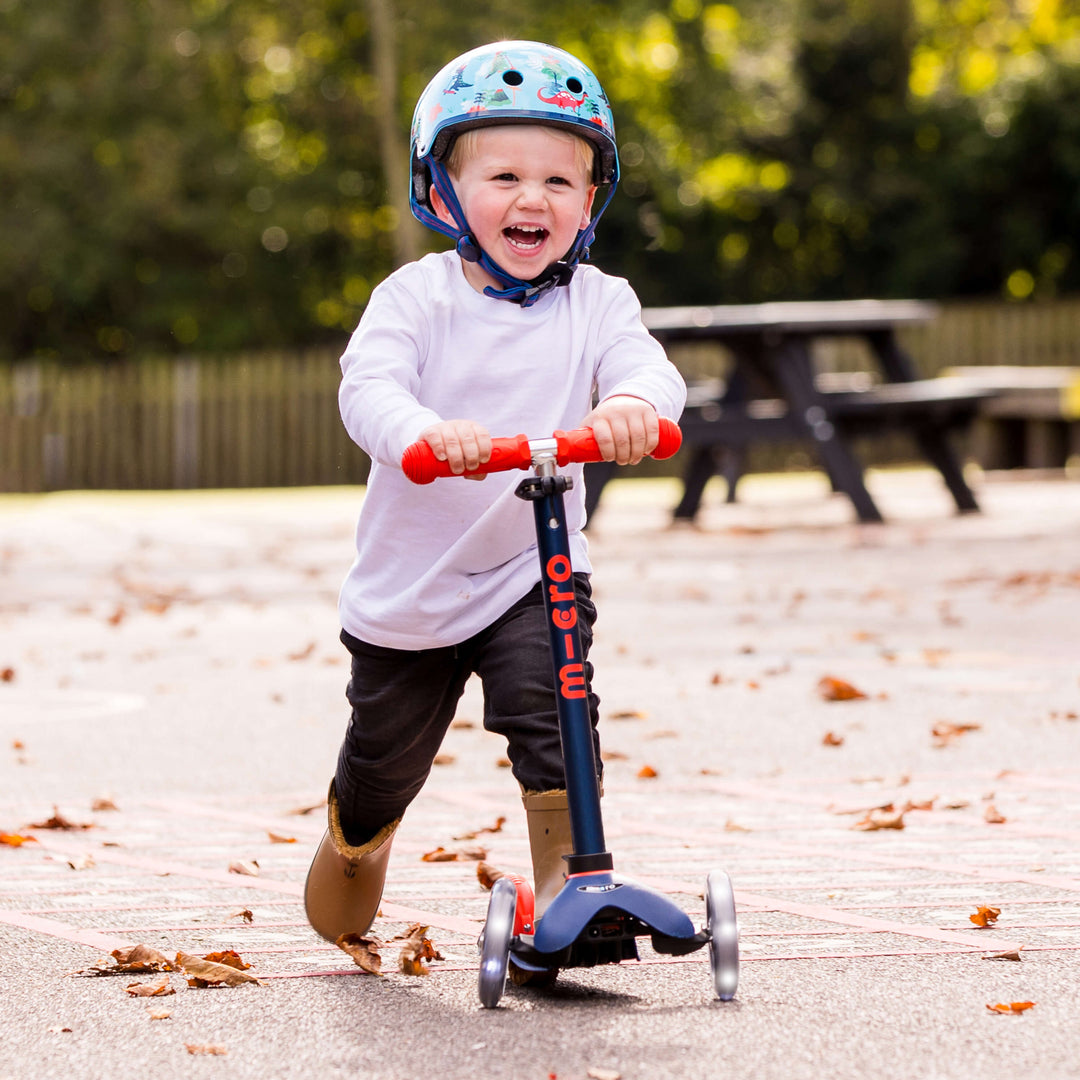 Laughing toddler in a helmet and white top riding the navy Mini Micro Deluxe scooter in a playground