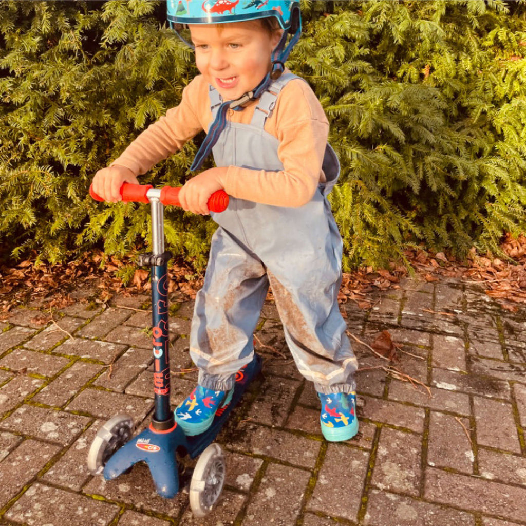 Smiling boy in a helmet and dungarees riding the navy Mini Micro Deluxe scooter on a brick path