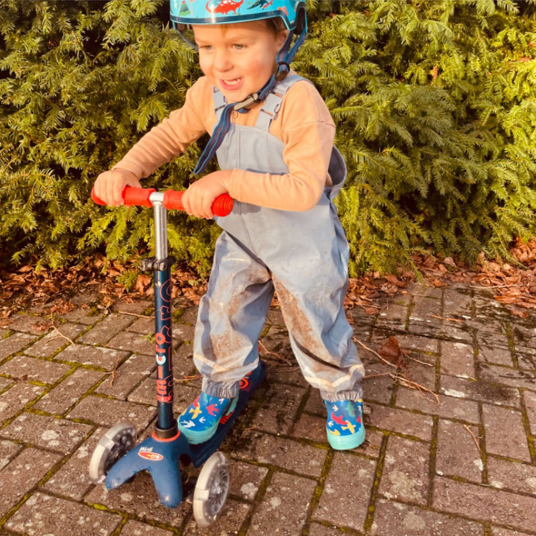 Smiling boy in a helmet and dungarees riding the navy Mini Micro Deluxe scooter on a brick path