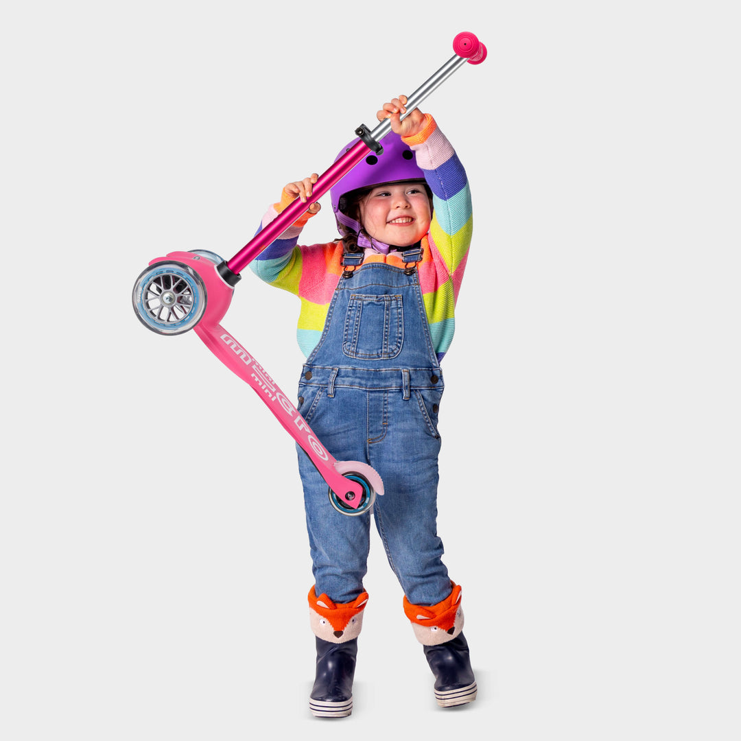 Toddler in a rainbow jumper and dungarees lifting the lightweight pink Mini Micro Deluxe scooter overhead