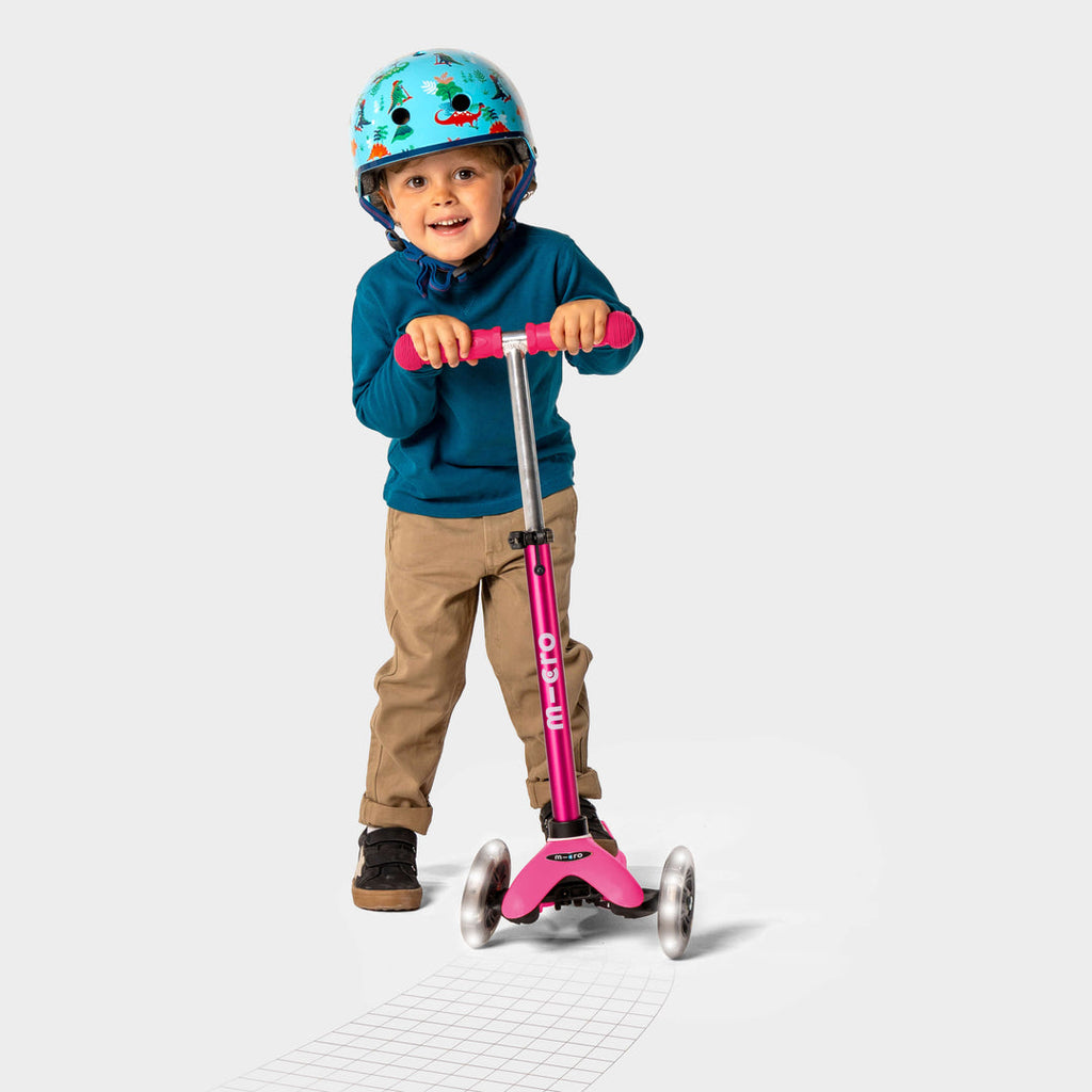 Smiling boy in a helmet and blue jumper riding the pink Mini Micro Deluxe LED scooter