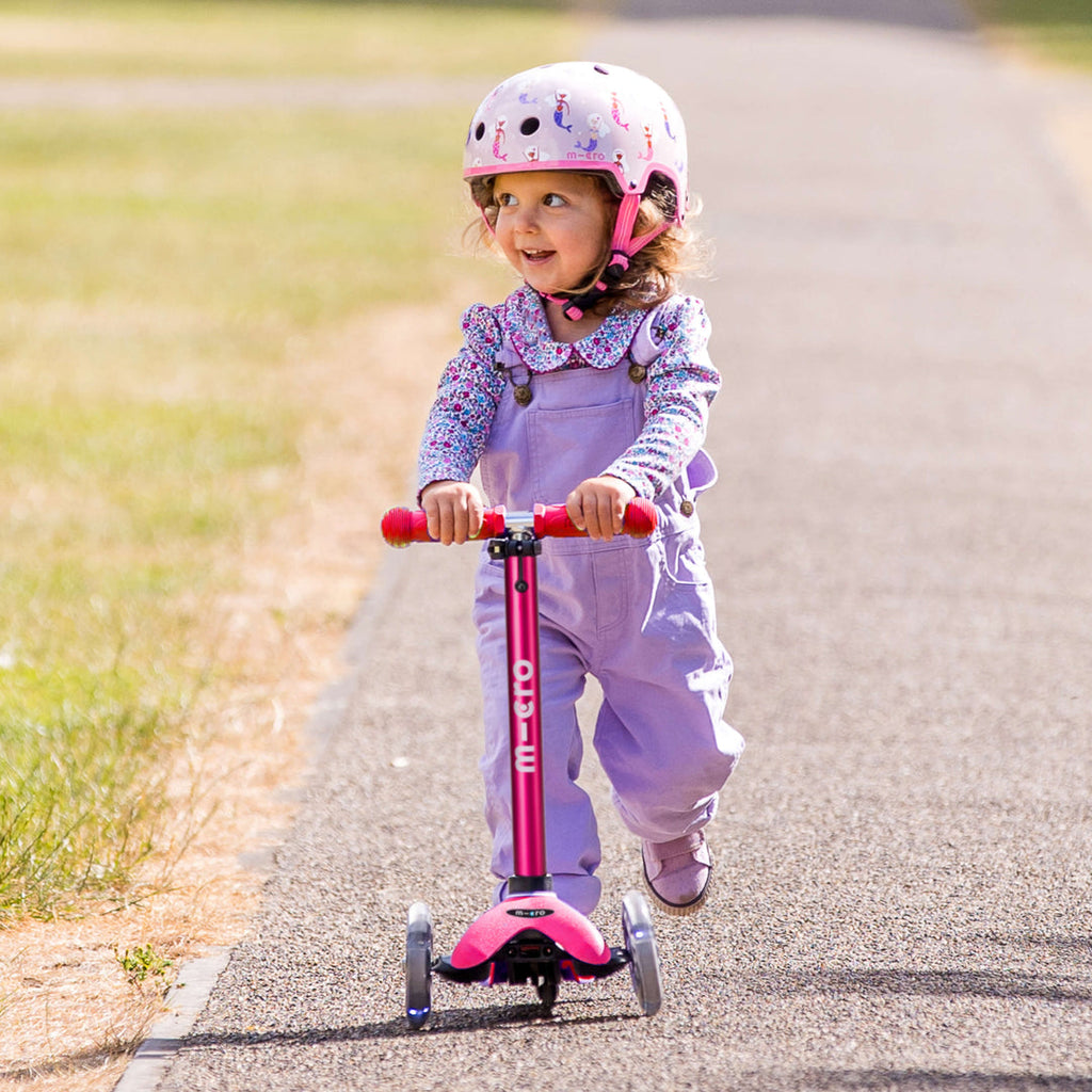 Toddler in a helmet and lilac dungarees riding the pink Mini Micro Deluxe scooter along a country path