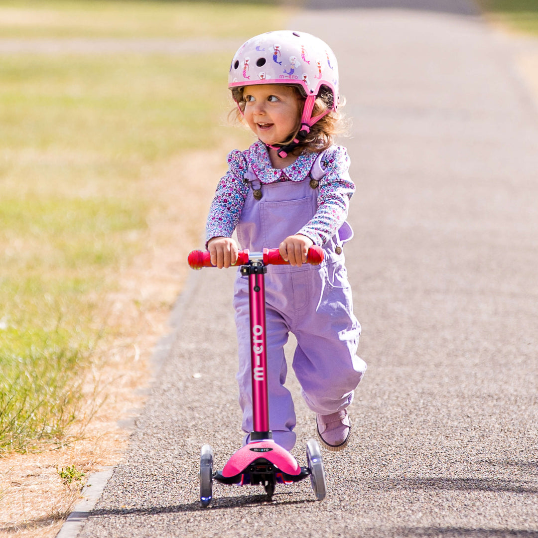Toddler in a helmet and lilac dungarees riding the pink Mini Micro Deluxe scooter along a country path