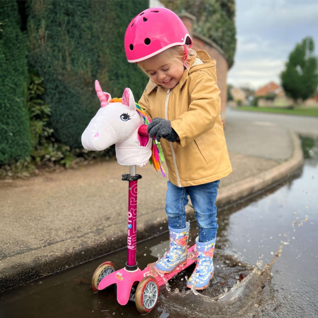 Toddler in a pink helmet and yellow raincoat splashing in a puddle on the pink Mini Micro scooter with a unicorn head