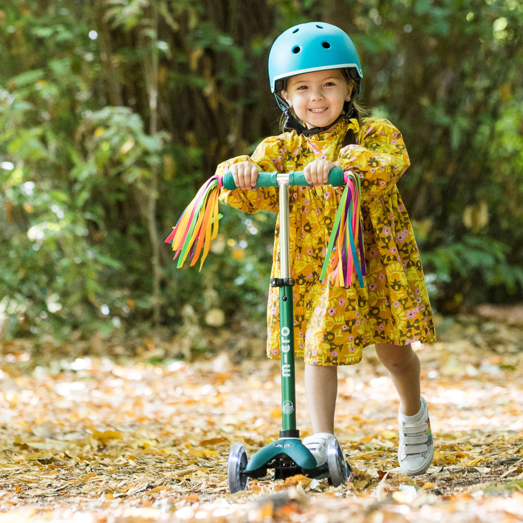 Smiling girl in a blue helmet and yellow floral dress riding the green Mini Micro Eco scooter with ribbon streamers in autumn leaves