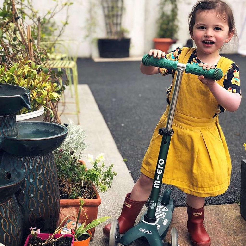 Smiling girl in a yellow pinafore and red boots standing with the green Mini Micro Eco scooter in a garden