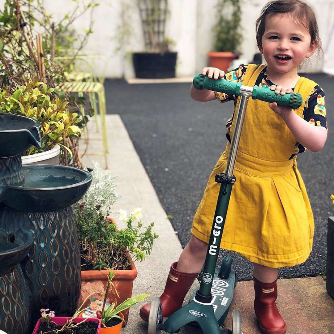 Smiling girl in a yellow pinafore and red boots standing with the green Mini Micro Eco scooter in a garden