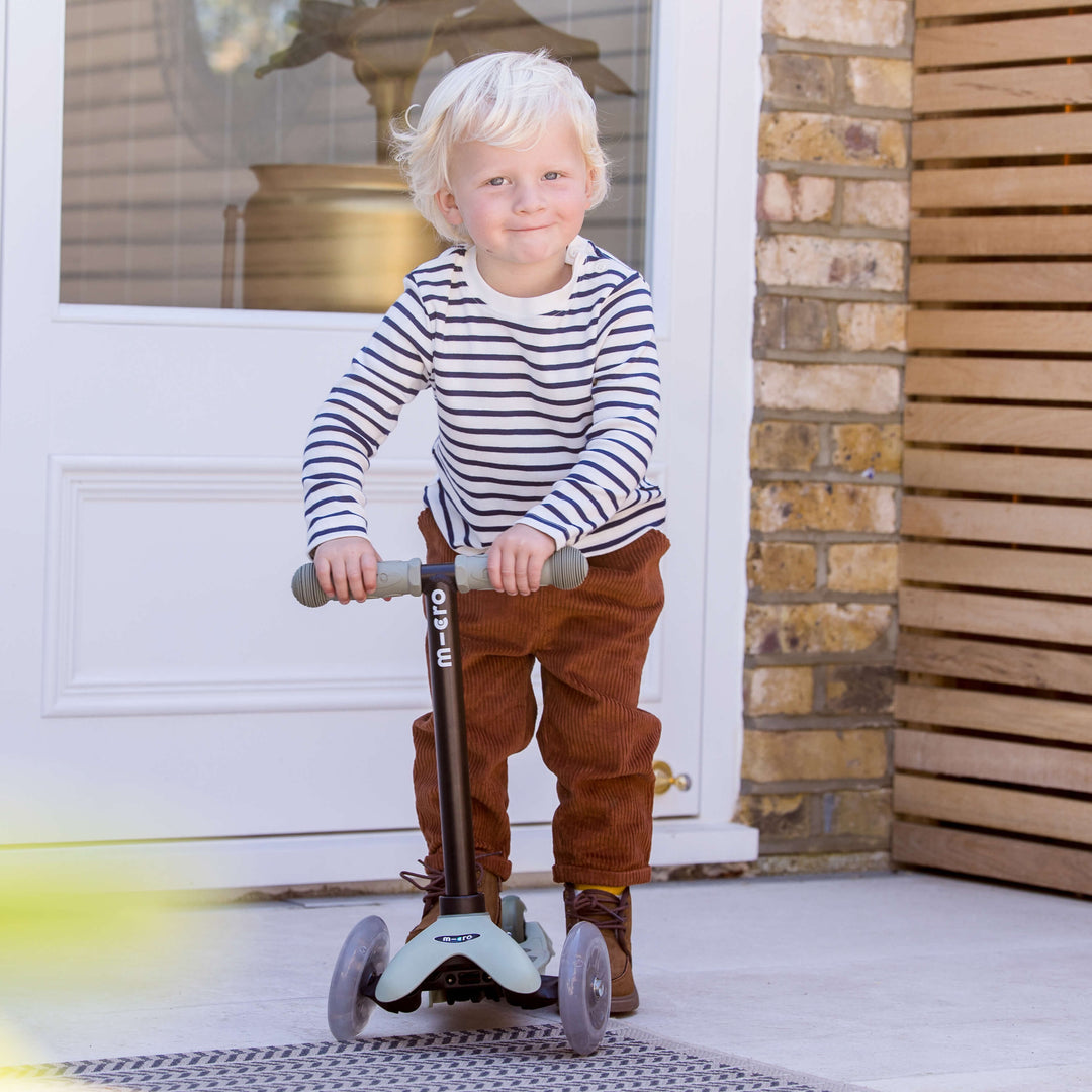 Toddler in a striped top riding the sage green Micro Mini2Grow Deluxe as a scooter outside a front door