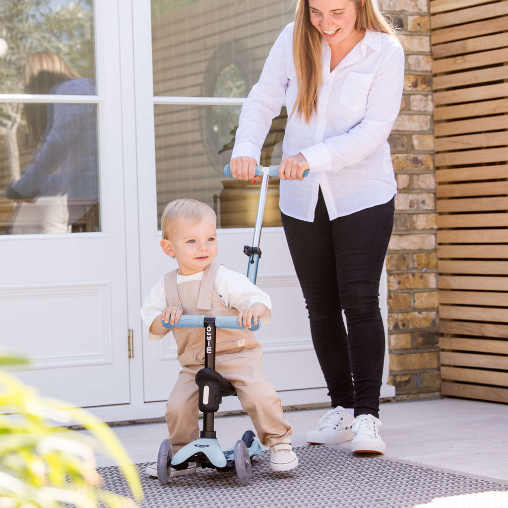 Parent pushing a baby on the soft blue Micro Mini2Grow Deluxe ride-on using the parent handle