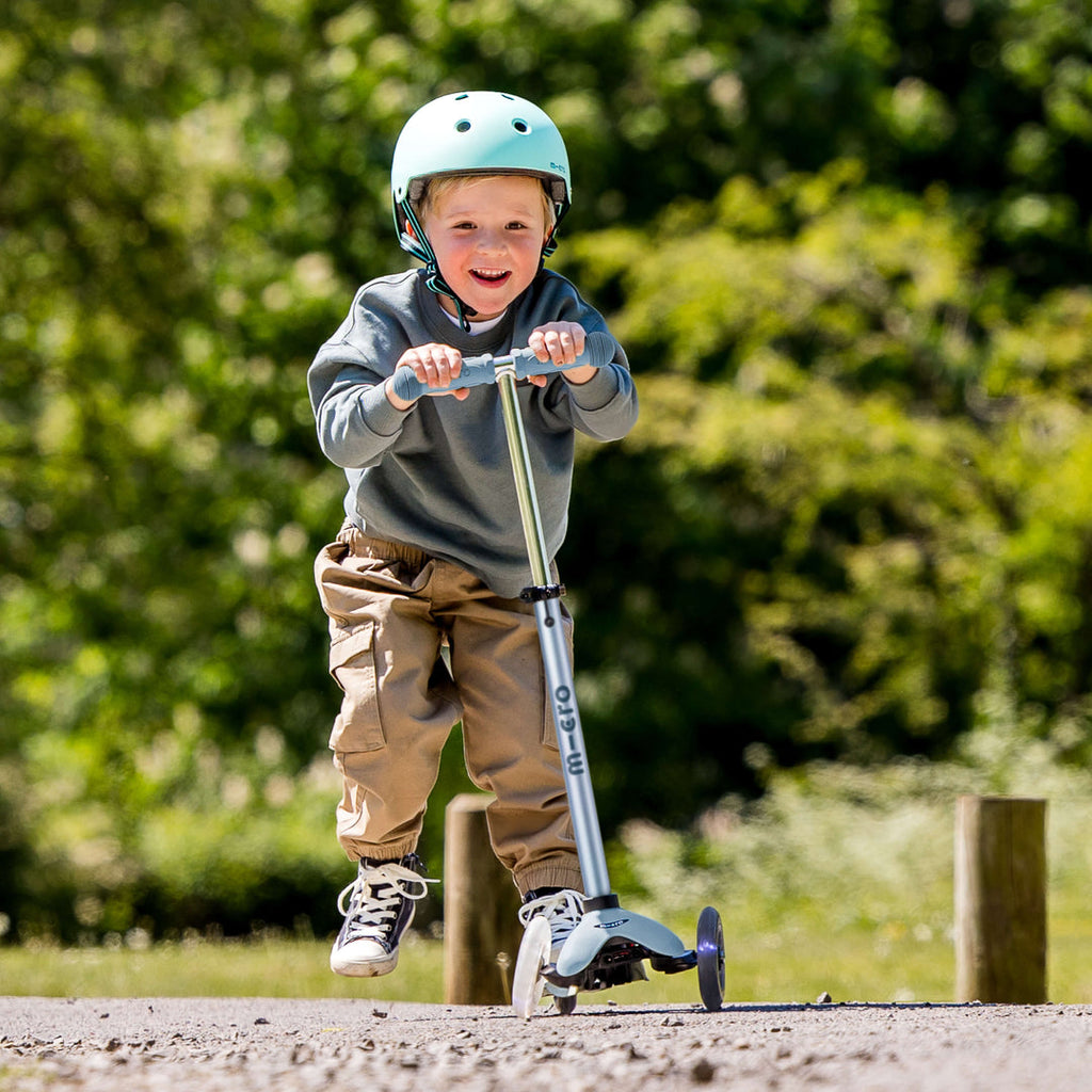 Smiling boy in a green helmet riding the soft blue Micro Mini2Grow Deluxe as a scooter on a park path
