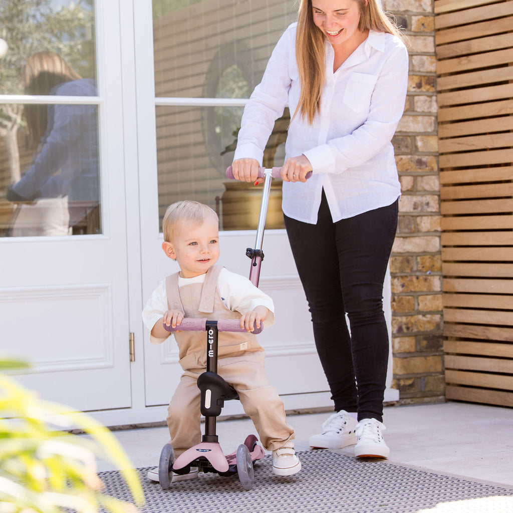 Parent pushing a baby on the rose pink Micro Mini2Grow Deluxe ride-on using the parent handle
