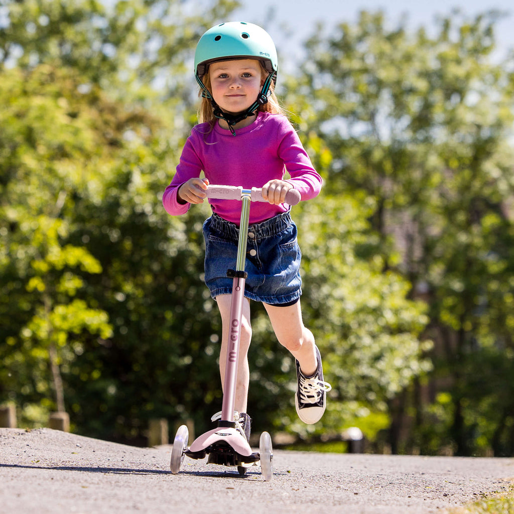 Girl in a helmet and pink top riding the rose pink Micro Mini2Grow Deluxe as a scooter on a park path