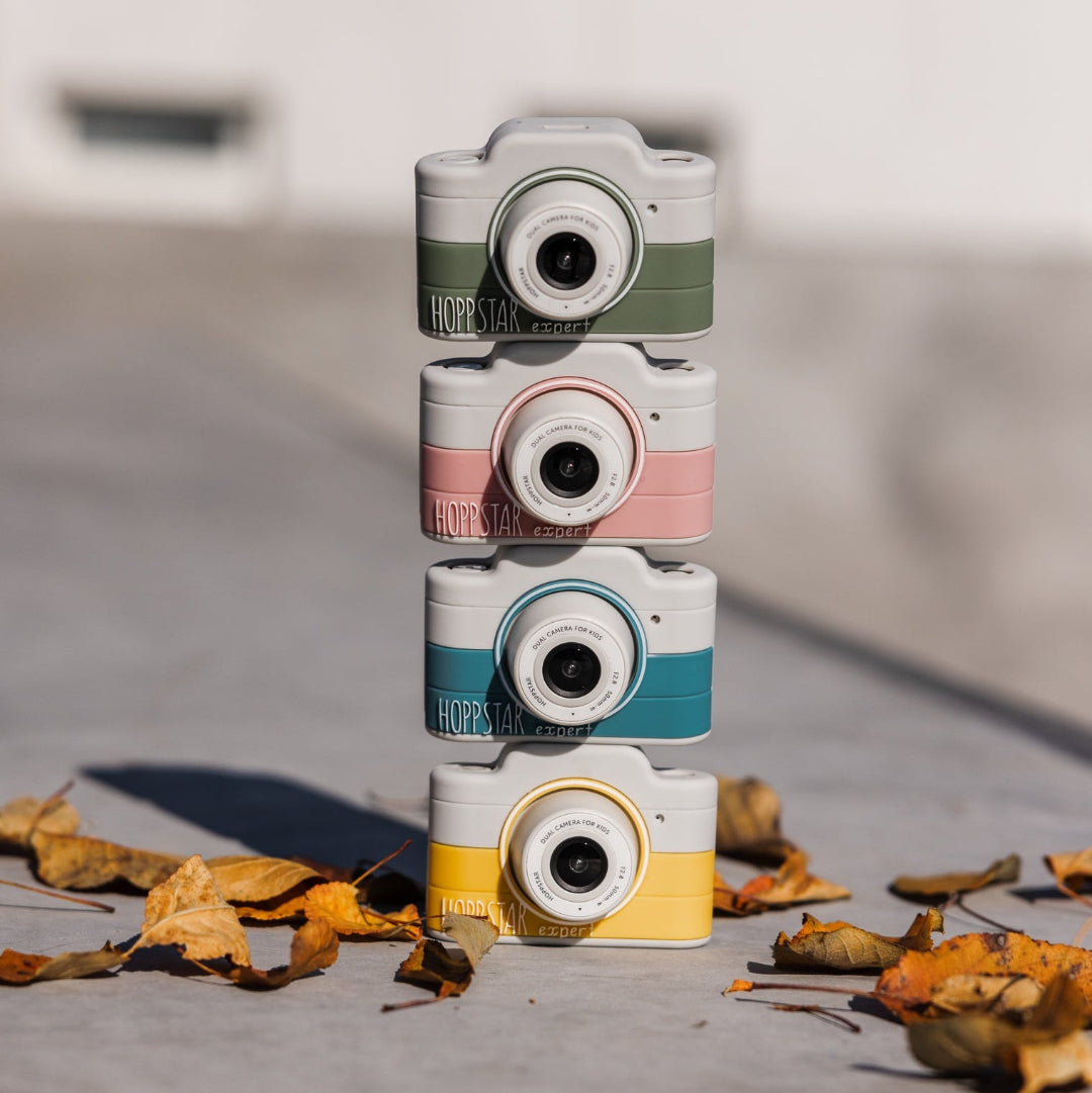 Four retro-style instant cameras stacked vertically on a concrete surface outdoors, each in a different pastel color: sage green, pink, teal, and yellow. Dry leaves are scattered around them, with a blurred building background.