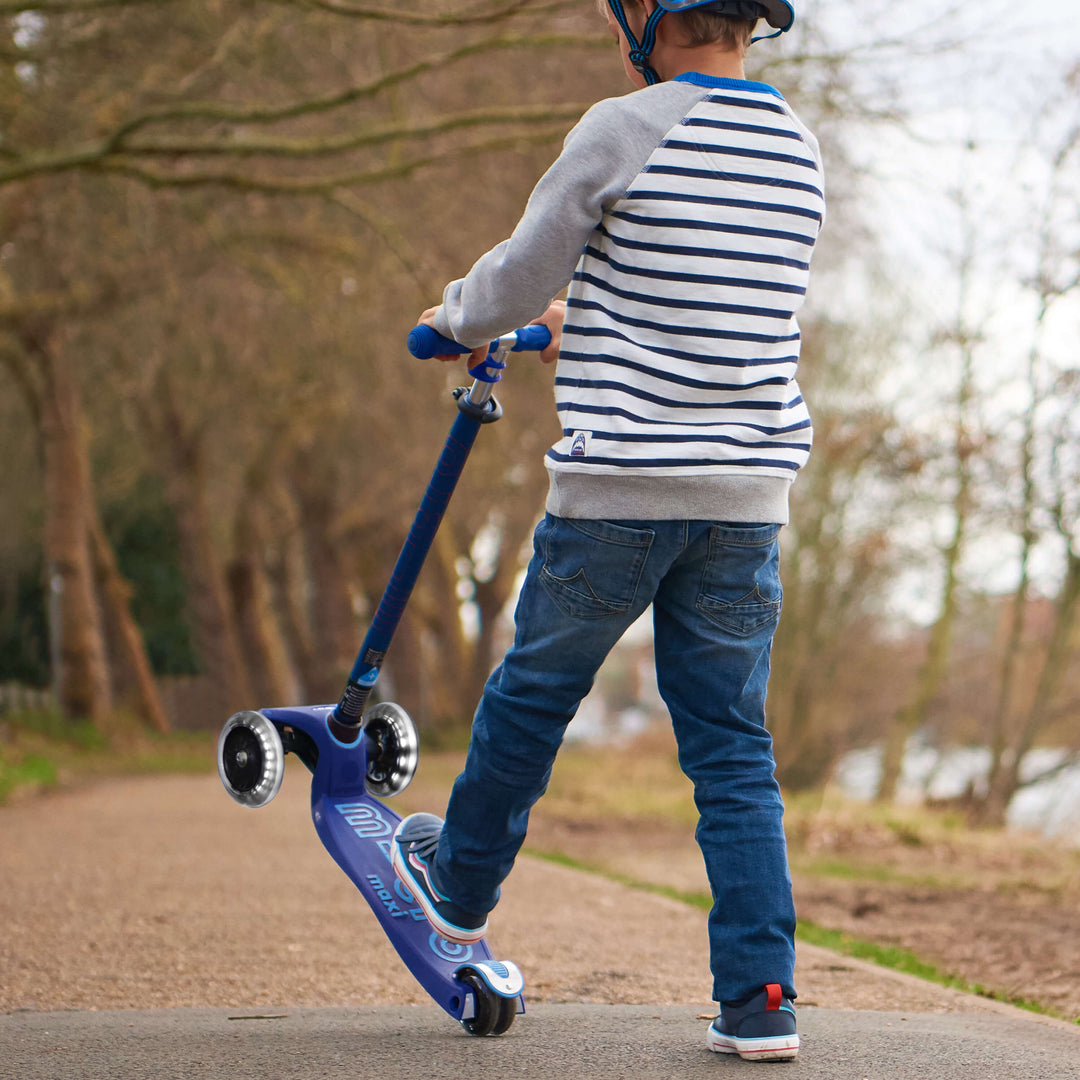 Child in a striped jumper riding the navy Maxi Micro Deluxe LED scooter along a woodland path