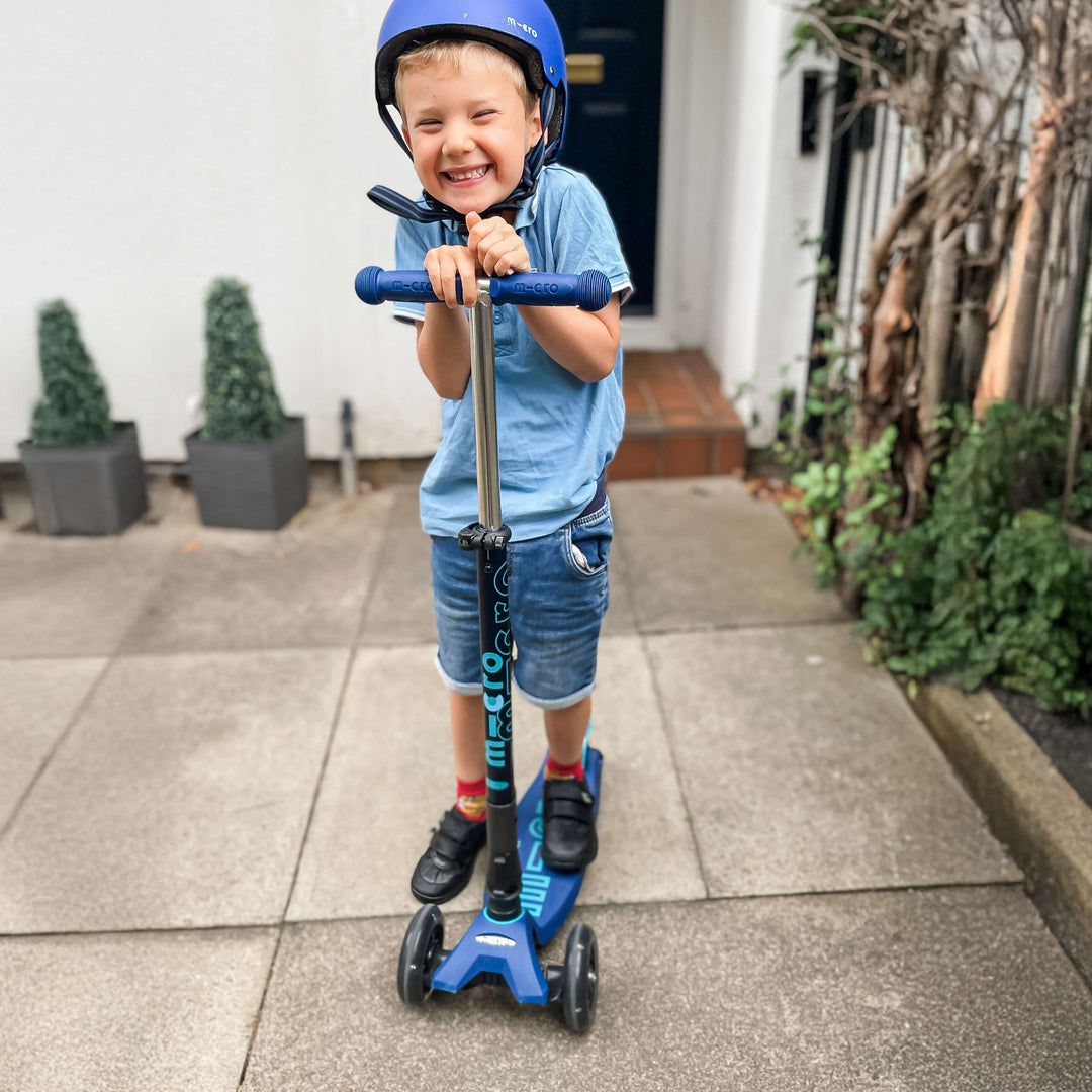 Smiling boy in a blue helmet holding the navy Maxi Micro Deluxe LED scooter outside a front door