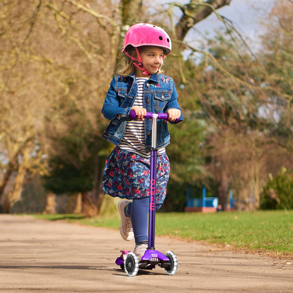 Smiling girl in a pink helmet and denim jacket riding the purple Maxi Micro Deluxe LED scooter in a park
