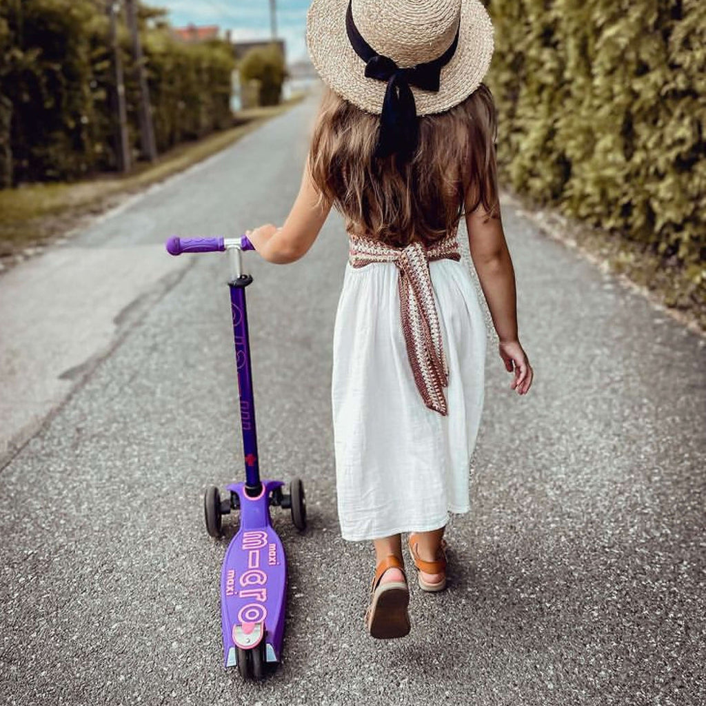 Girl in a sun hat and white dress walking down a lane pulling the purple Maxi Micro Deluxe scooter