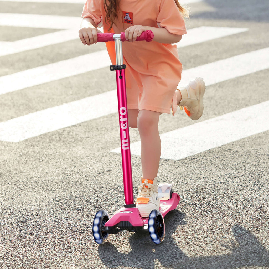 Girl in an orange outfit riding the pink Maxi Micro Deluxe LED scooter across a zebra crossing with the wheels lit up