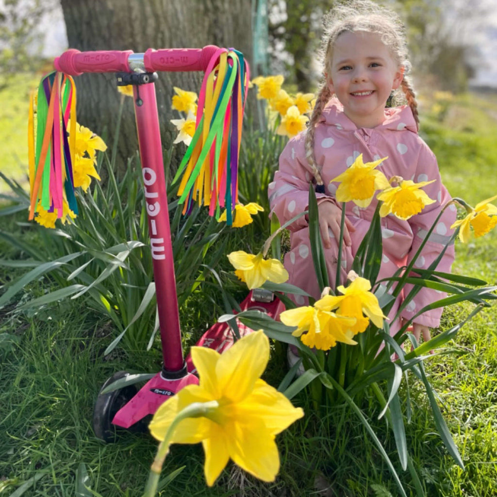 Smiling girl in a pink raincoat among daffodils next to the pink Maxi Micro Deluxe scooter with rainbow handlebar ribbons