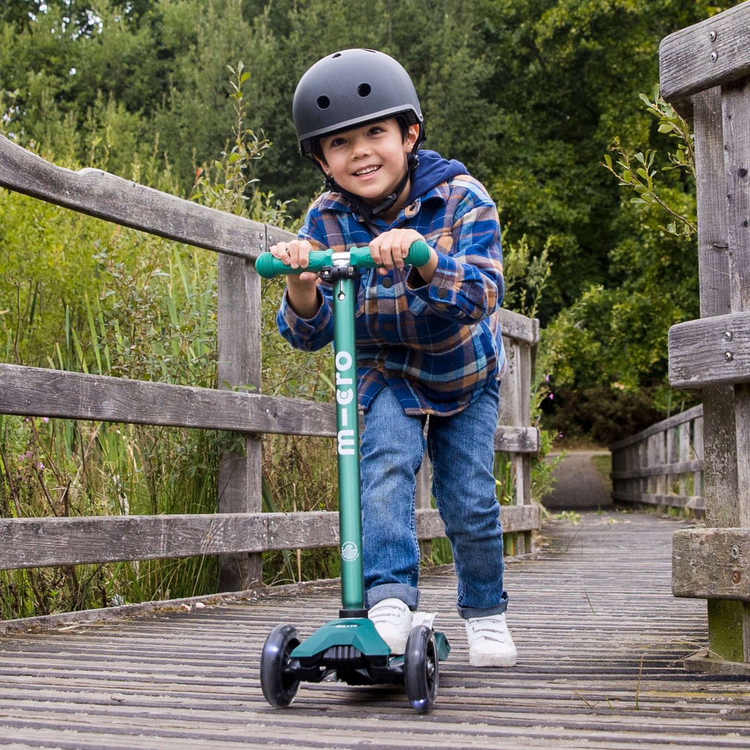 Smiling boy in a helmet and checked shirt riding the green Maxi Micro Eco scooter on a wooden bridge