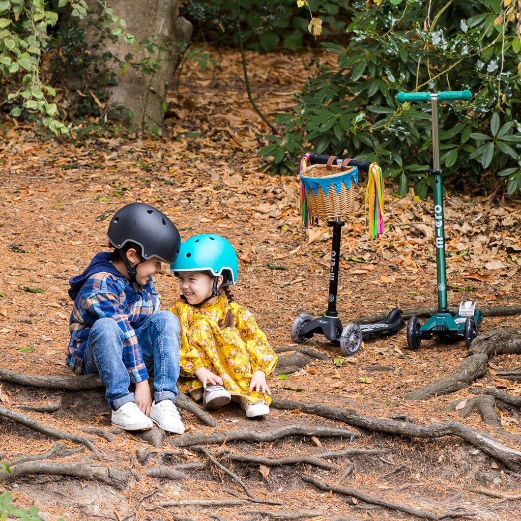 Two children in helmets sitting on tree roots in a forest beside their Micro scooters