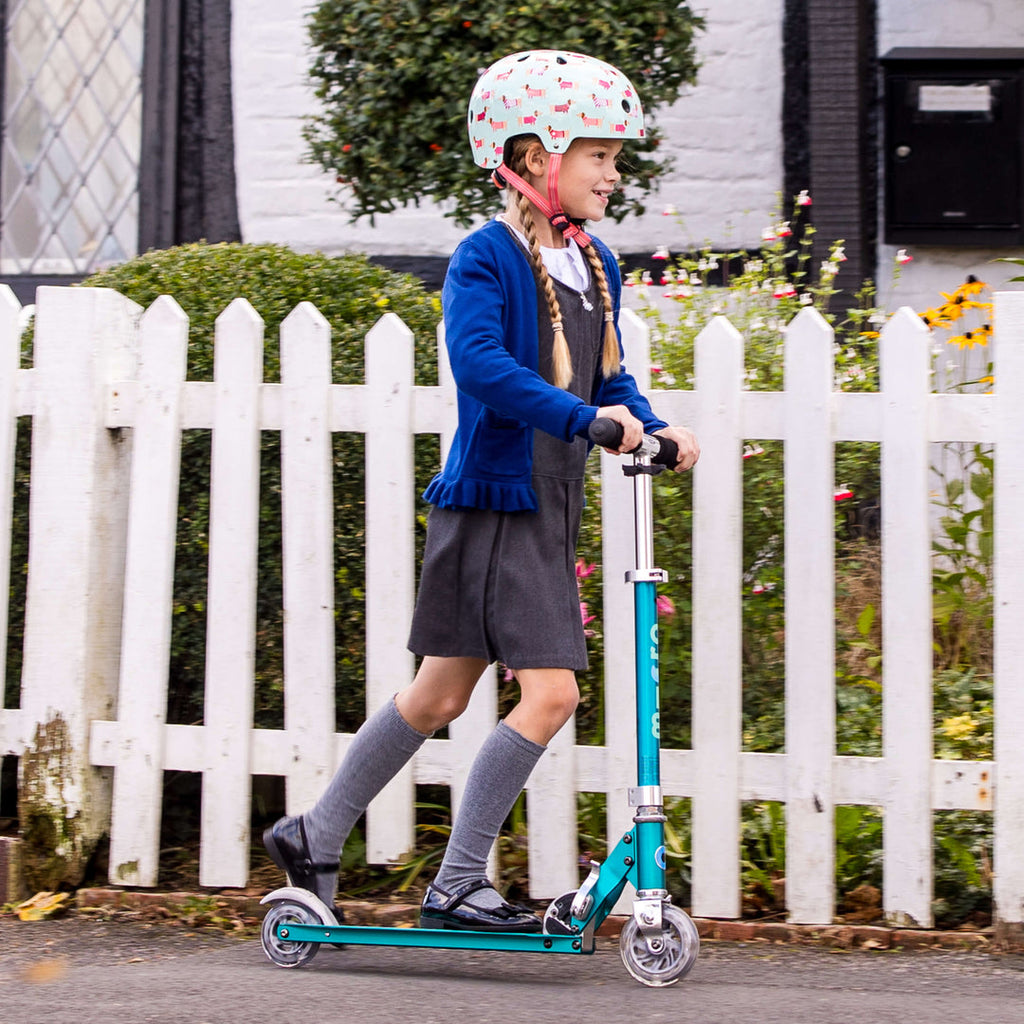 Girl in a school uniform and helmet riding the aqua Micro Sprite LED scooter past a white picket fence
