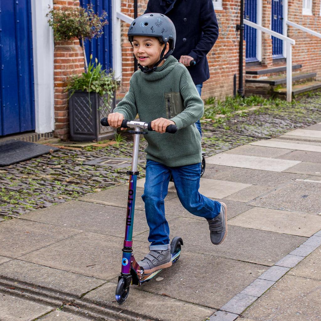 Smiling boy in a helmet and green hoodie riding the Neochrome Micro Sprite LED scooter along a pavement