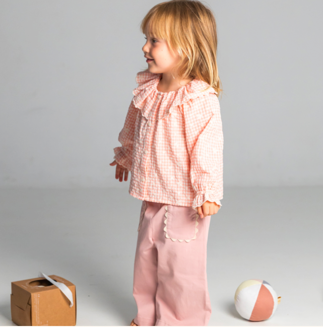 Young girl in pink checkered top and pants standing on a gray background with a cardboard box and ball.